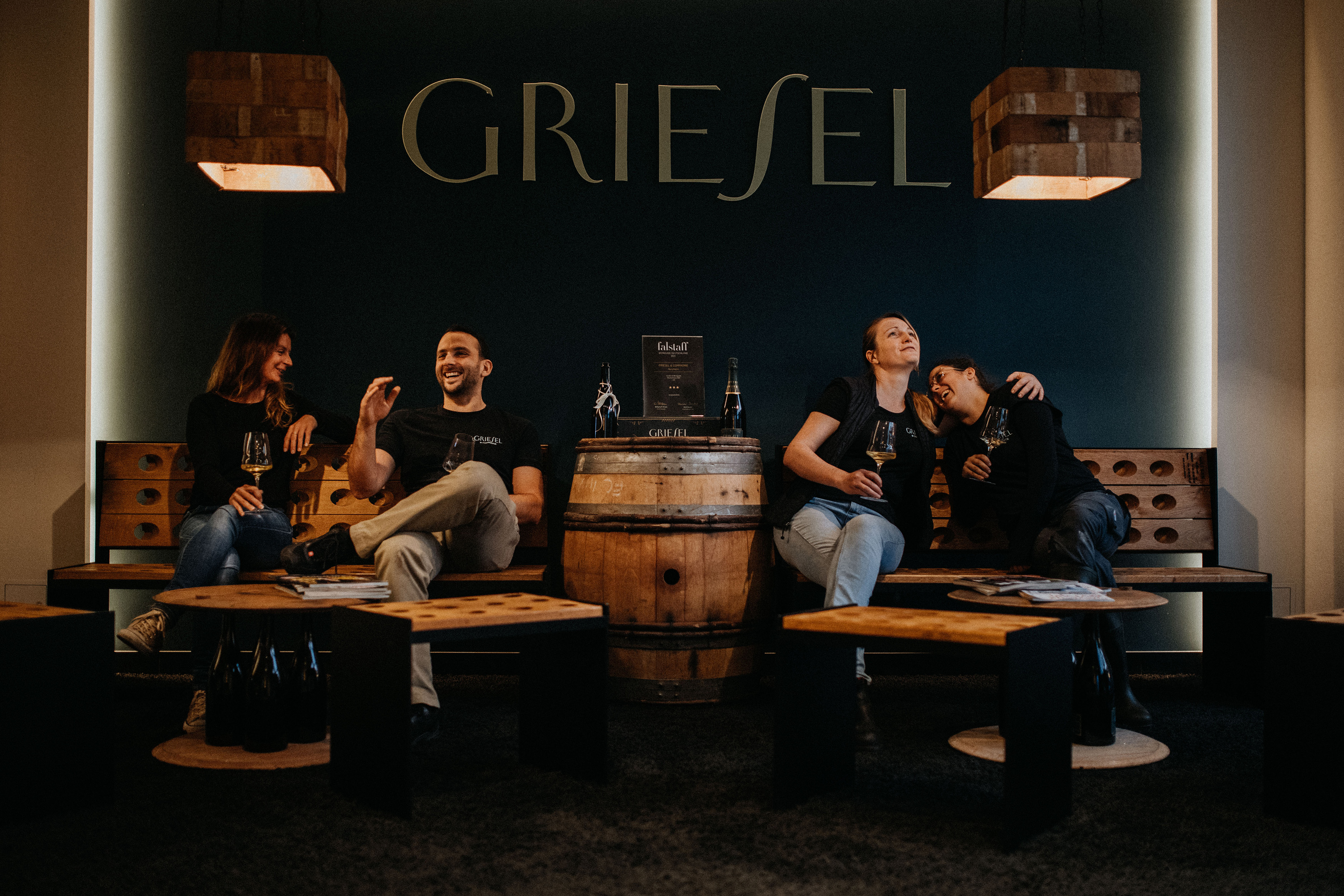 4 Personen vom Team Griesel sitzen auf einer Bank mit Tischen und trinken ein Glas Sekt und lachen