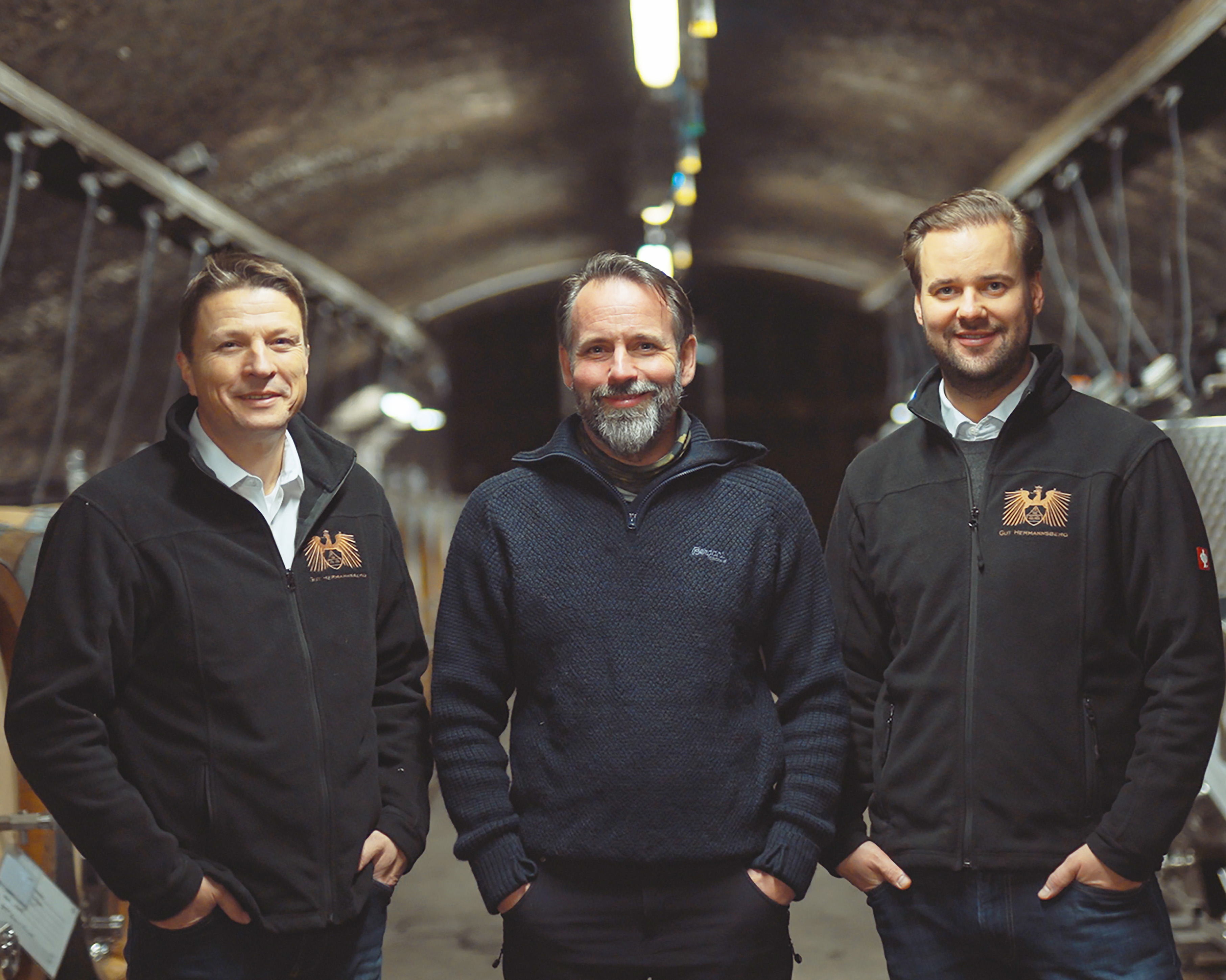 Drei Männer vom Gut Hermannsbergstehen im Weinkeller und lächeln in die Kamera.